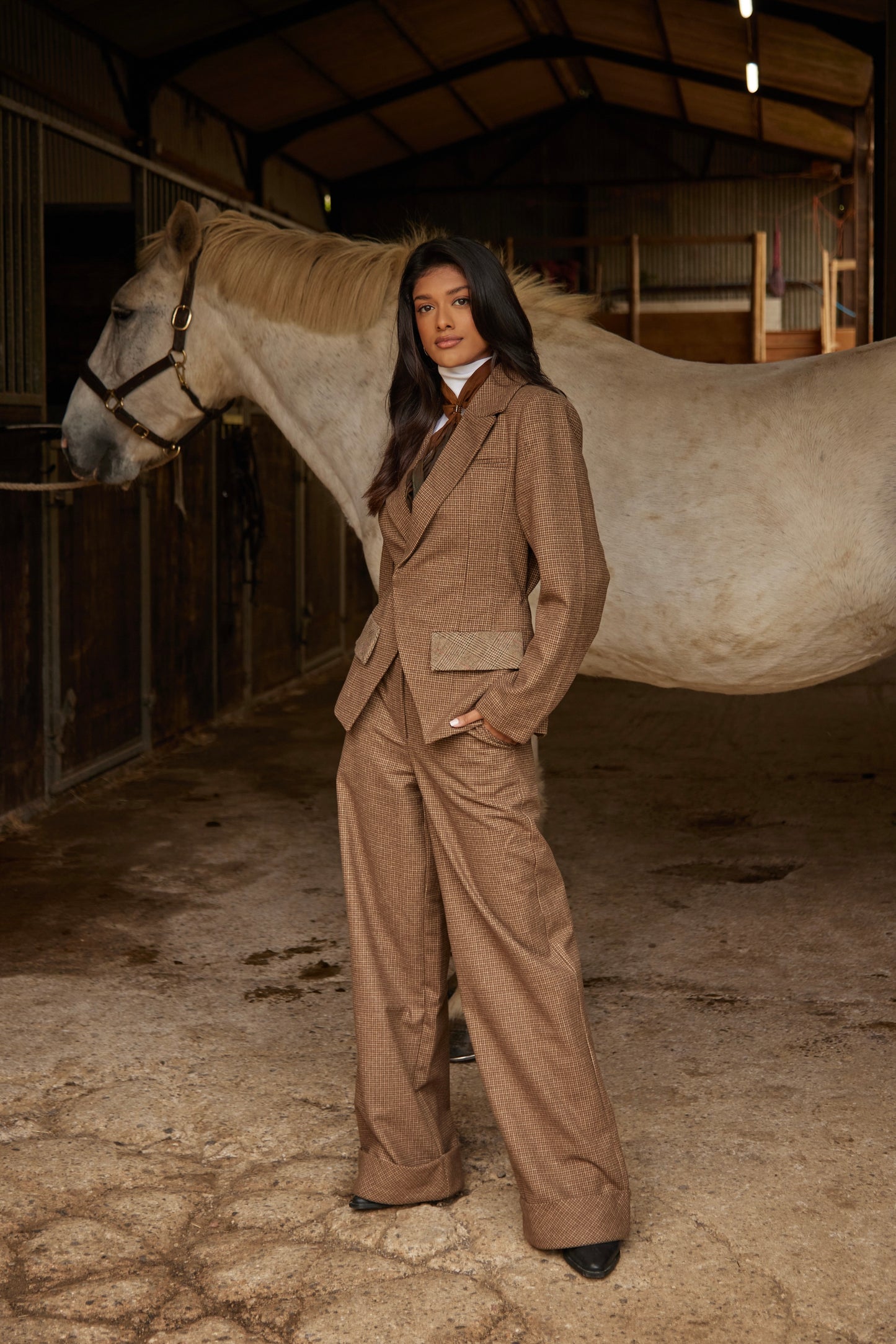 Woman in a beige suit standing next to a horse in a stable
