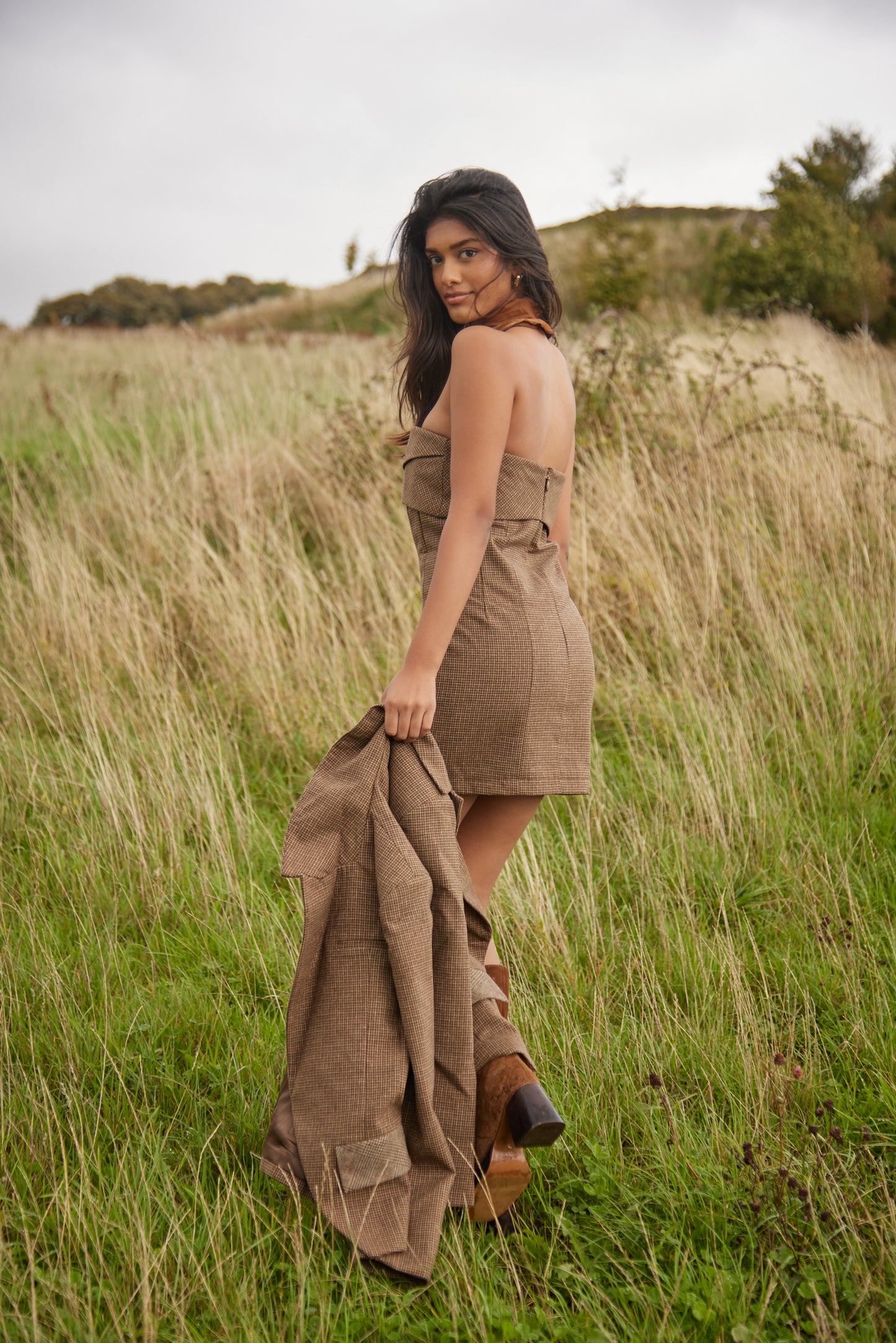 Woman in a brown dress holding a matching jacket in a grassy field
