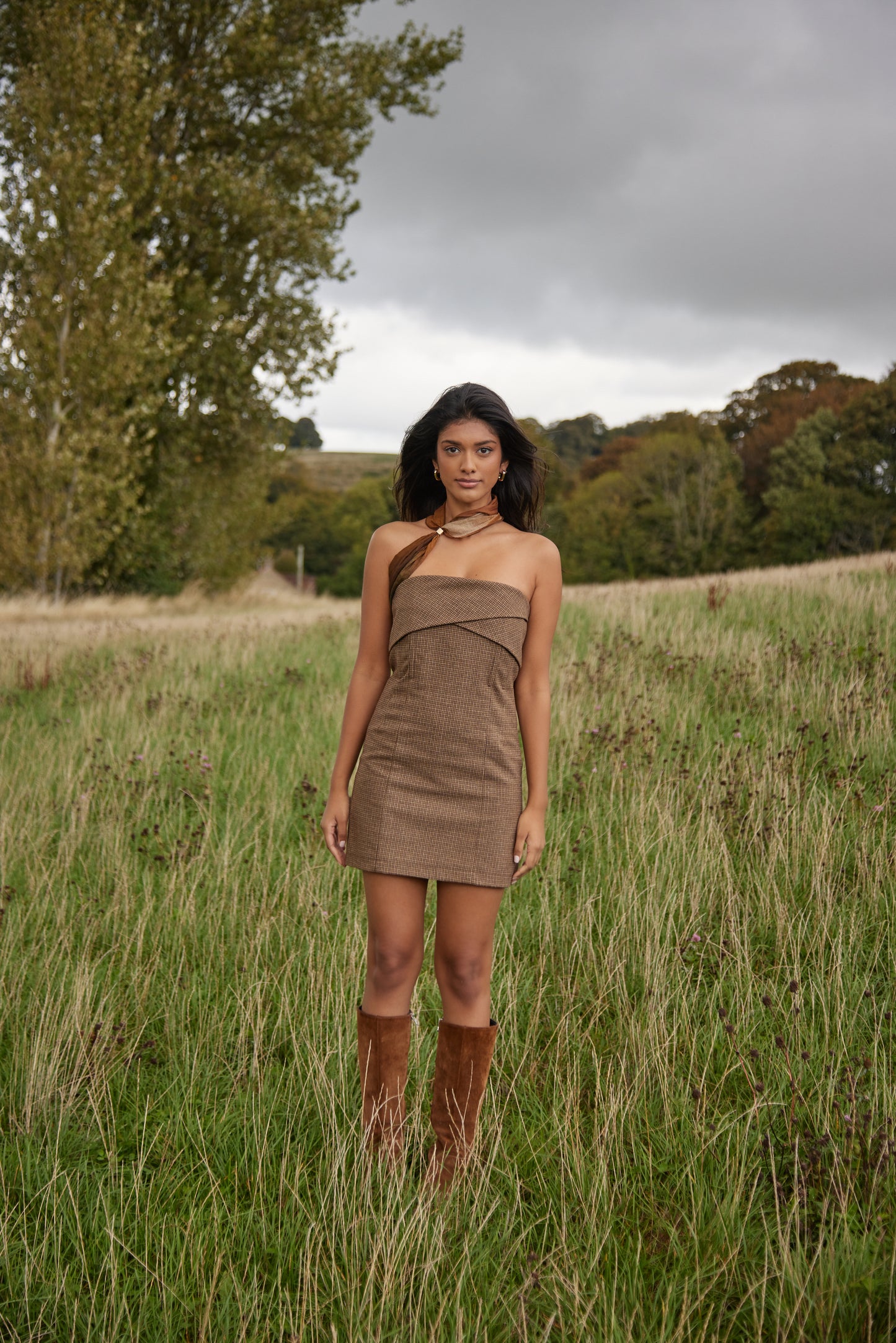 Woman in a brown dress standing in a grassy field with trees in the background