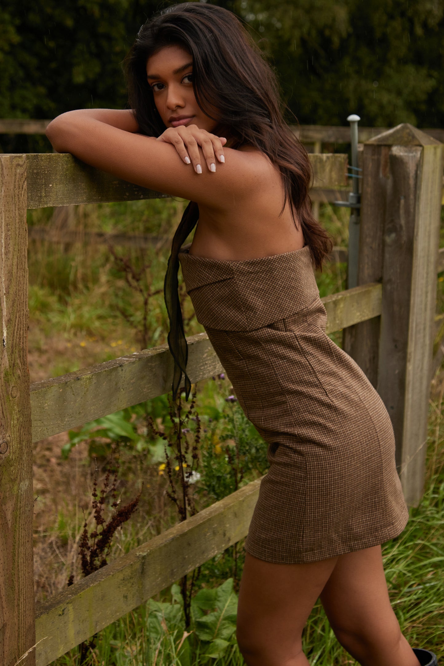 Woman in a brown dress leaning against a wooden fence in a natural setting