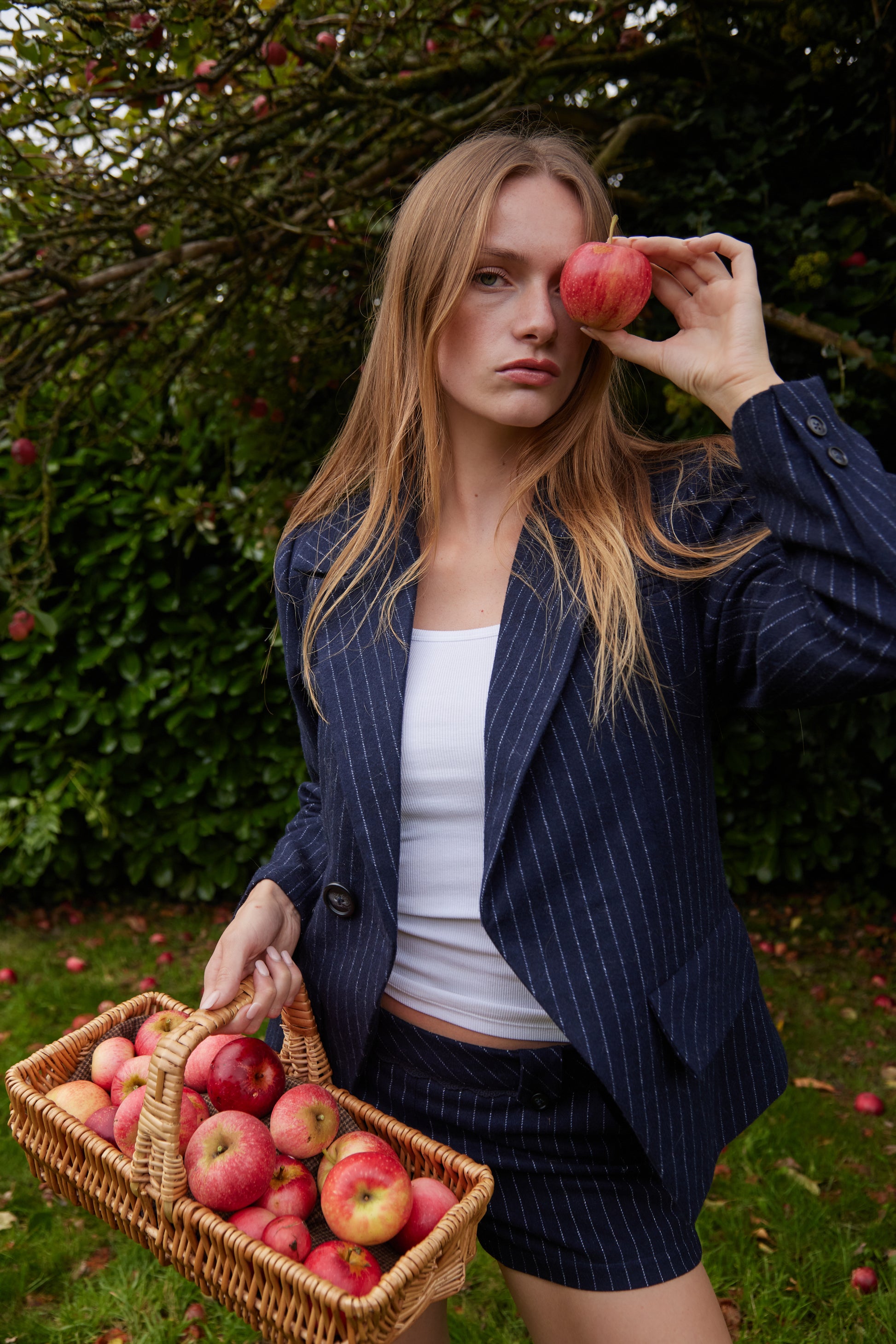 Woman holding a basket of apples and an apple to her eye in an orchard.